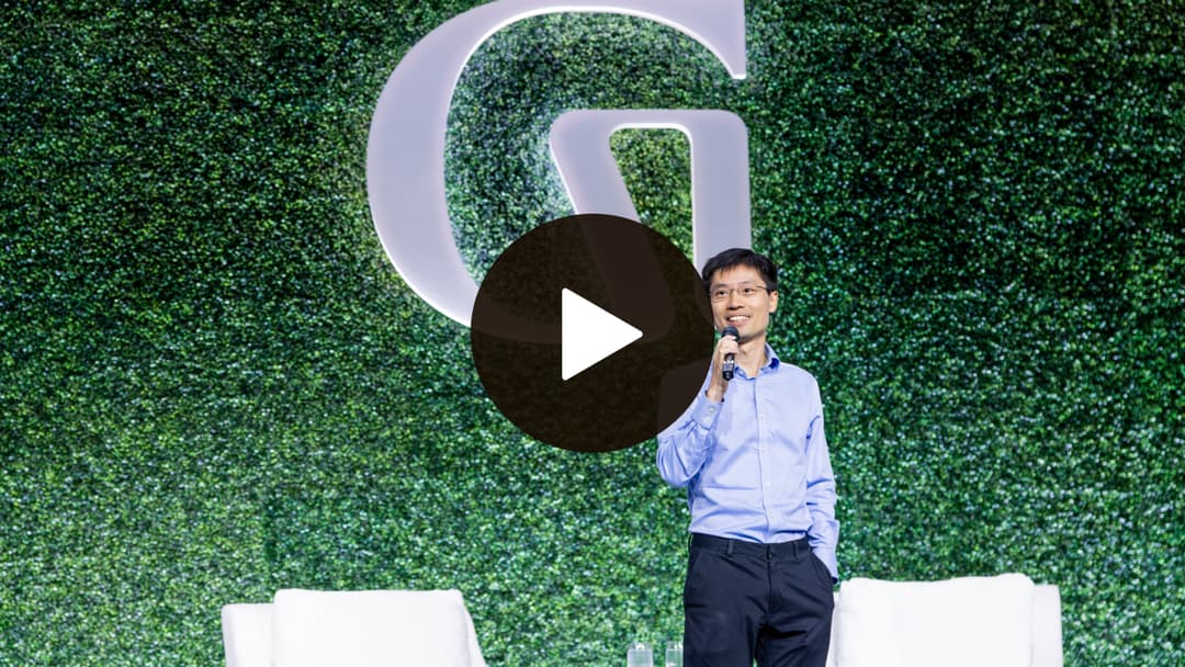 Man with a microphone stands on stage with a large "G" logo behind, set against a green leafy background. Two white chairs are visible.