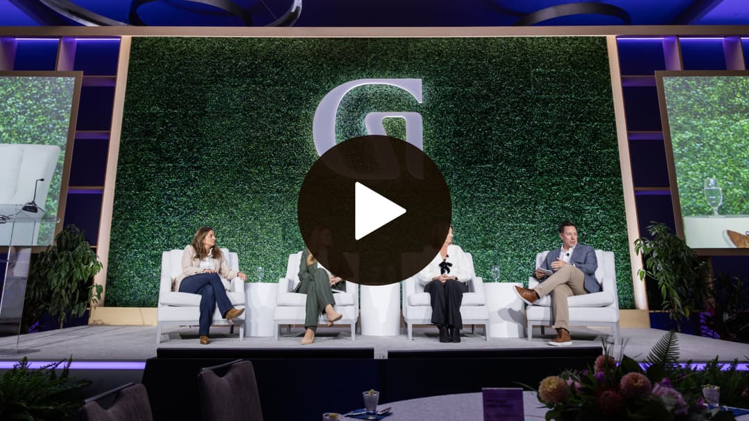A panel of four people seated on stage with green foliage backdrop and large "G" logo, surrounded by screens displaying the discussion.